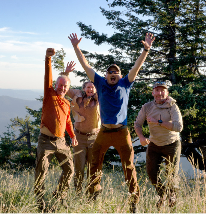 Group of four people in field clothes jumping with excitement on grassy ridge top with pine tree in background